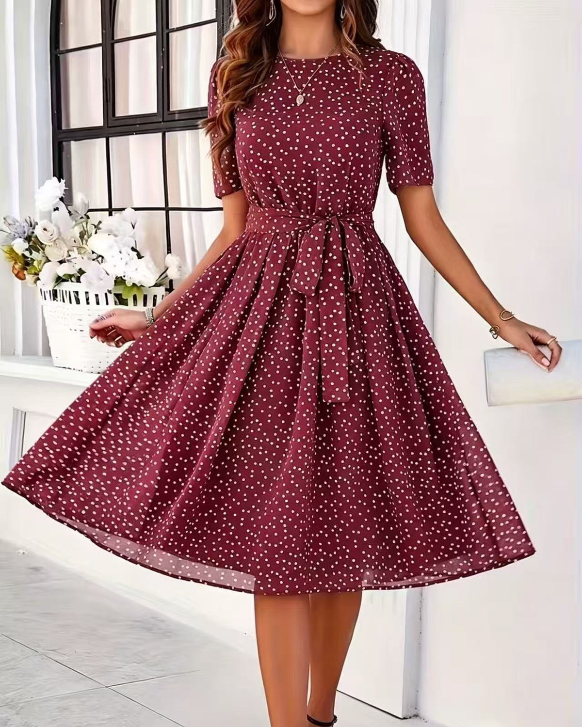 A woman wearing a short-sleeve, knee-length A-line dress in burgundy with a white polka dot pattern. The dress features a round neckline, a self-tie waist belt, and a flared skirt.
