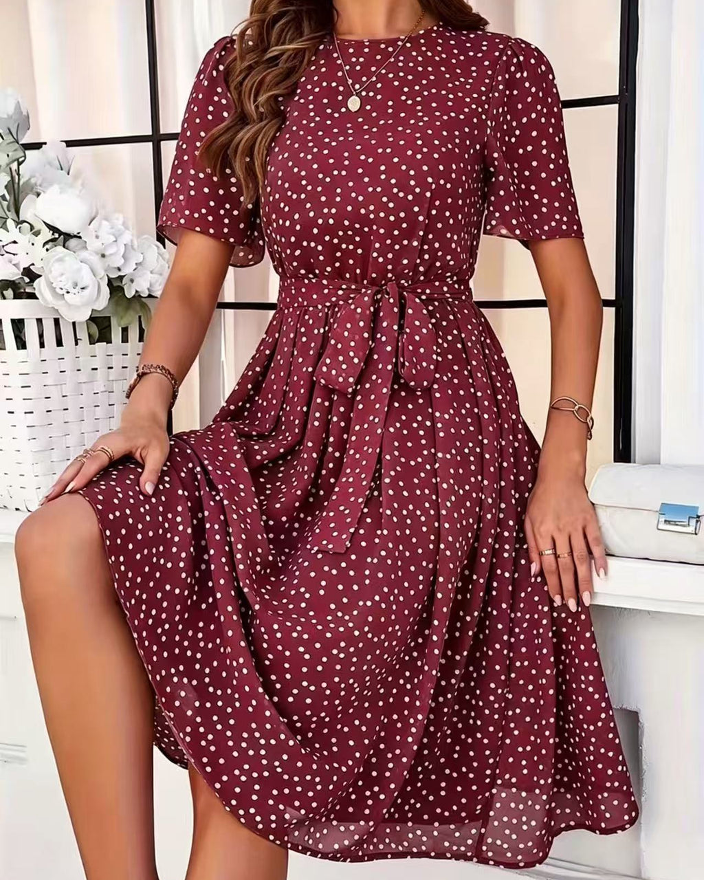 A woman wearing a short-sleeve midi dress with a red base and white polka dot pattern, sitting in front of a white window frame.