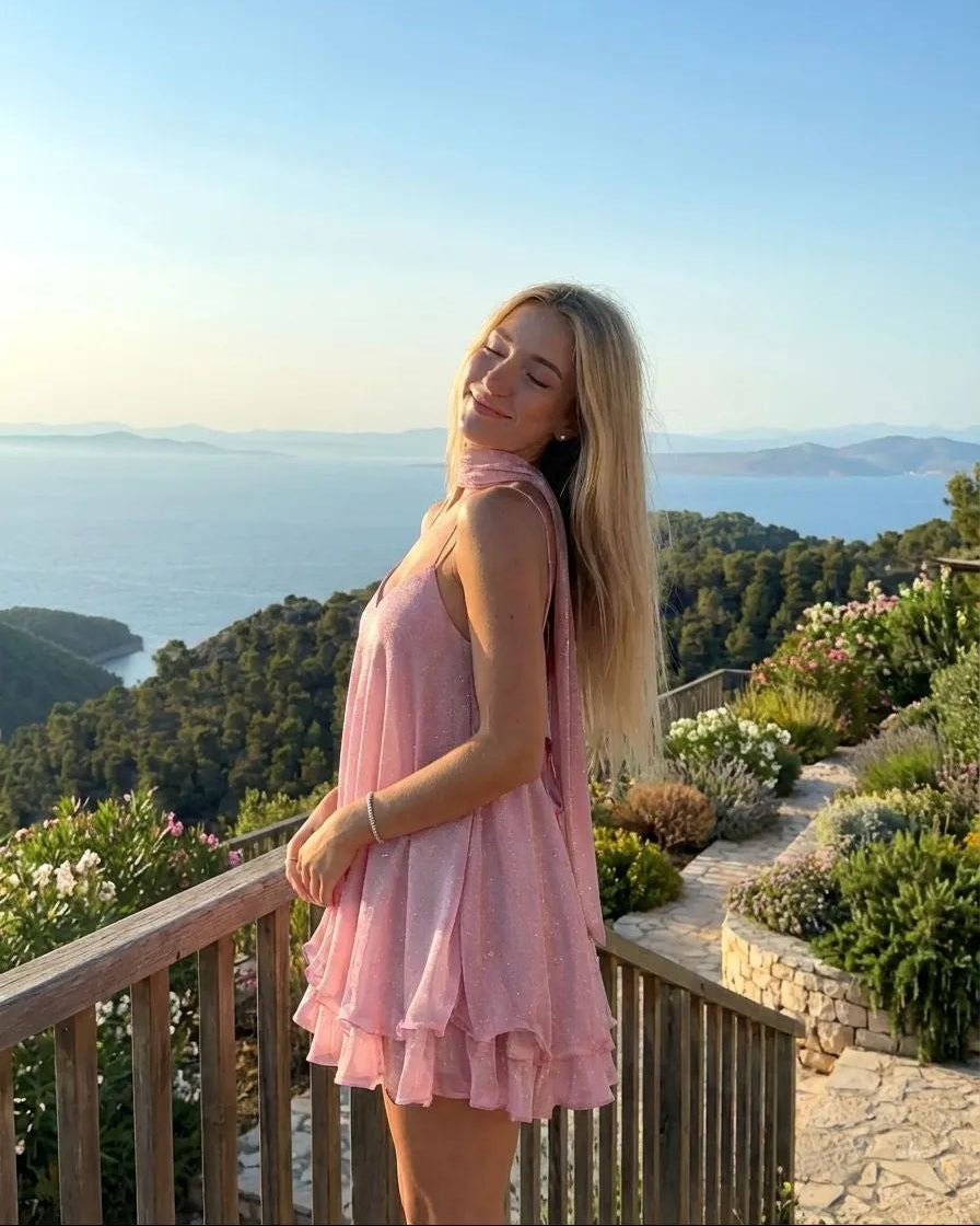 Woman in a pink dress standing on a balcony with a scenic view of the sea and mountains.
