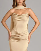 A woman modeling a champagne beige satin halter-neck gown with structured corset-style draping and gathered details, photographed against a solid gray studio background.