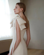 A woman wearing a sleeveless cream-colored dress with a large bow detail on the shoulder, standing in a softly lit indoor setting.