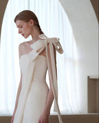 A woman wearing a cream-colored one-shoulder evening gown with a large bow detail on the back, standing in a softly lit interior with an arched window background.
