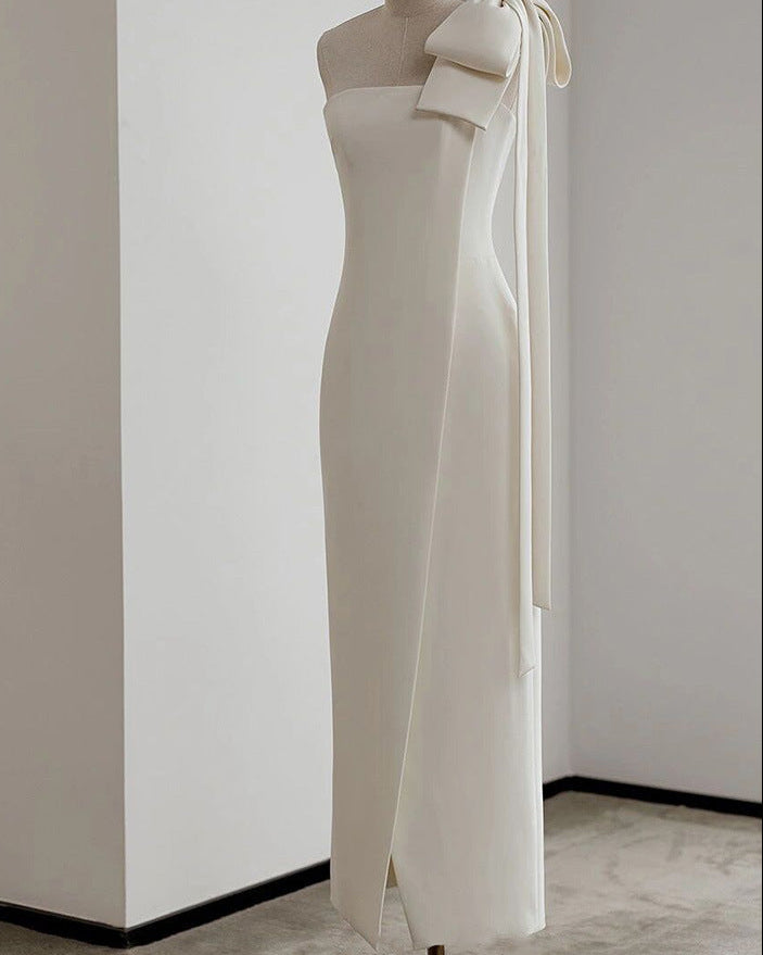 An off-white, one-shoulder midi dress with a large bow detail on the shoulder and a long tie ribbon, displayed on a mannequin in a minimalist room with white walls and a gray floor.
