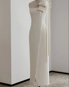 An off-white, one-shoulder midi dress with a large bow detail on the shoulder and a long tie ribbon, displayed on a mannequin in a minimalist room with white walls and a gray floor.