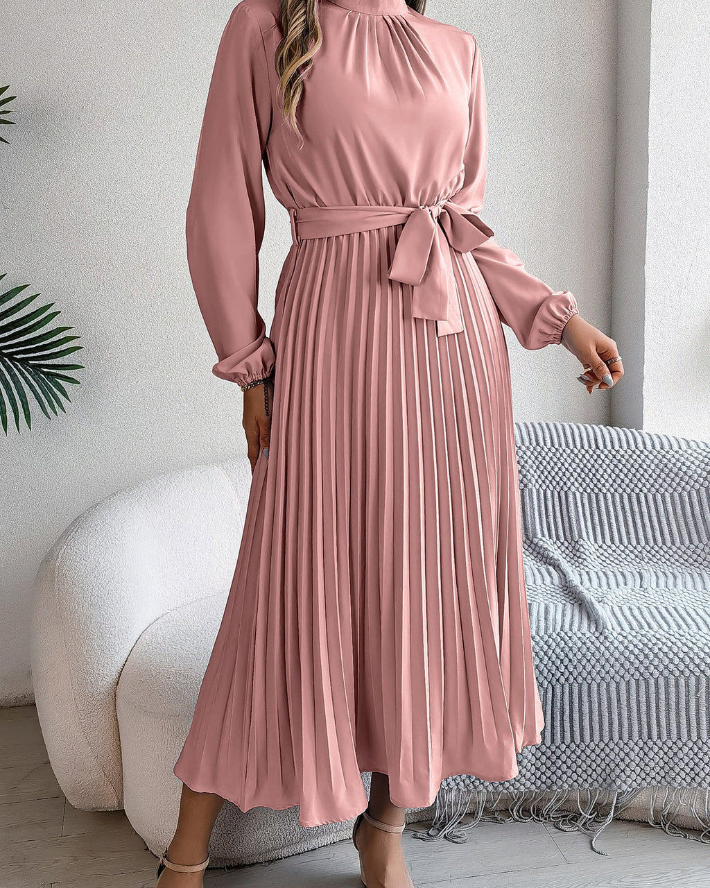 Woman wearing a pink pleated dress in a room with a white couch and decorative plant.