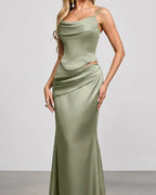 A woman models a floor-length sage green satin gown with spaghetti straps, a draped cowl neckline, a fitted waist, and a ruched skirt, set against a neutral studio background.