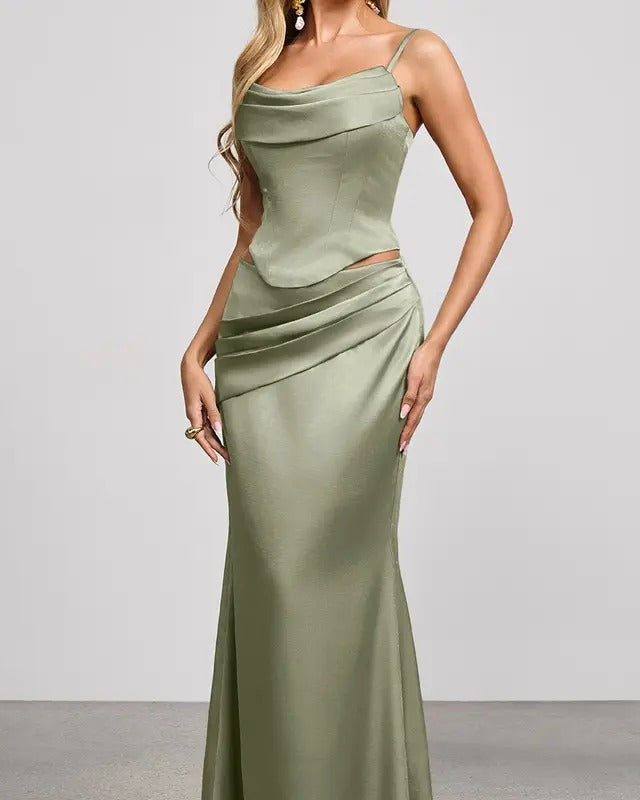 A woman models a floor-length sage green satin gown with spaghetti straps, a draped cowl neckline, a fitted waist, and a ruched skirt, set against a neutral studio background.