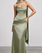 A woman modeling a floor-length sage green satin gown with a draped halter-style neckline, structured bodice, and asymmetrical draped skirt detail against a plain light gray studio background.