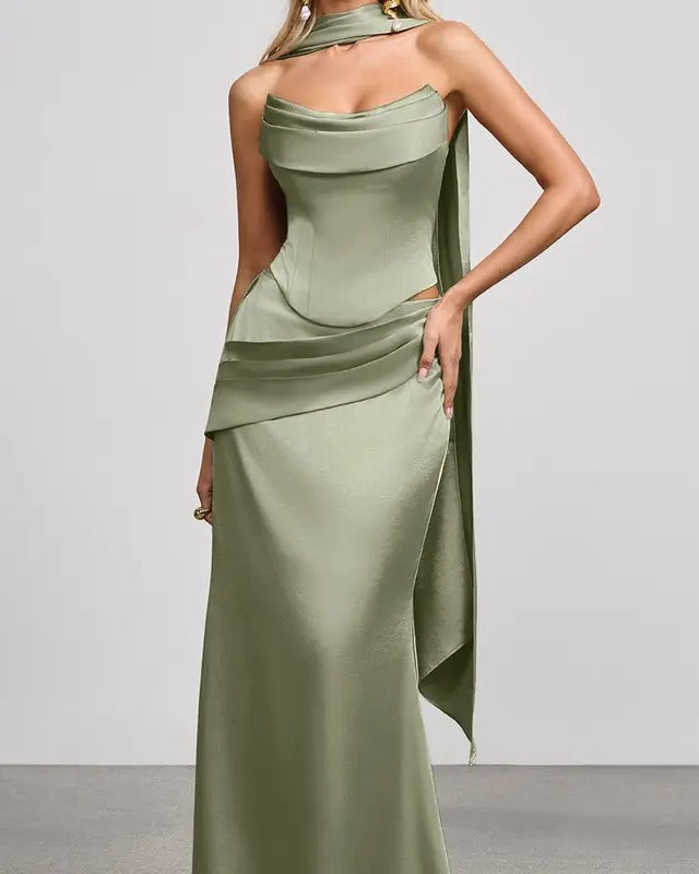 A woman modeling a floor-length sage green satin gown with a draped halter-style neckline, structured bodice, and asymmetrical draped skirt detail against a plain light gray studio background.