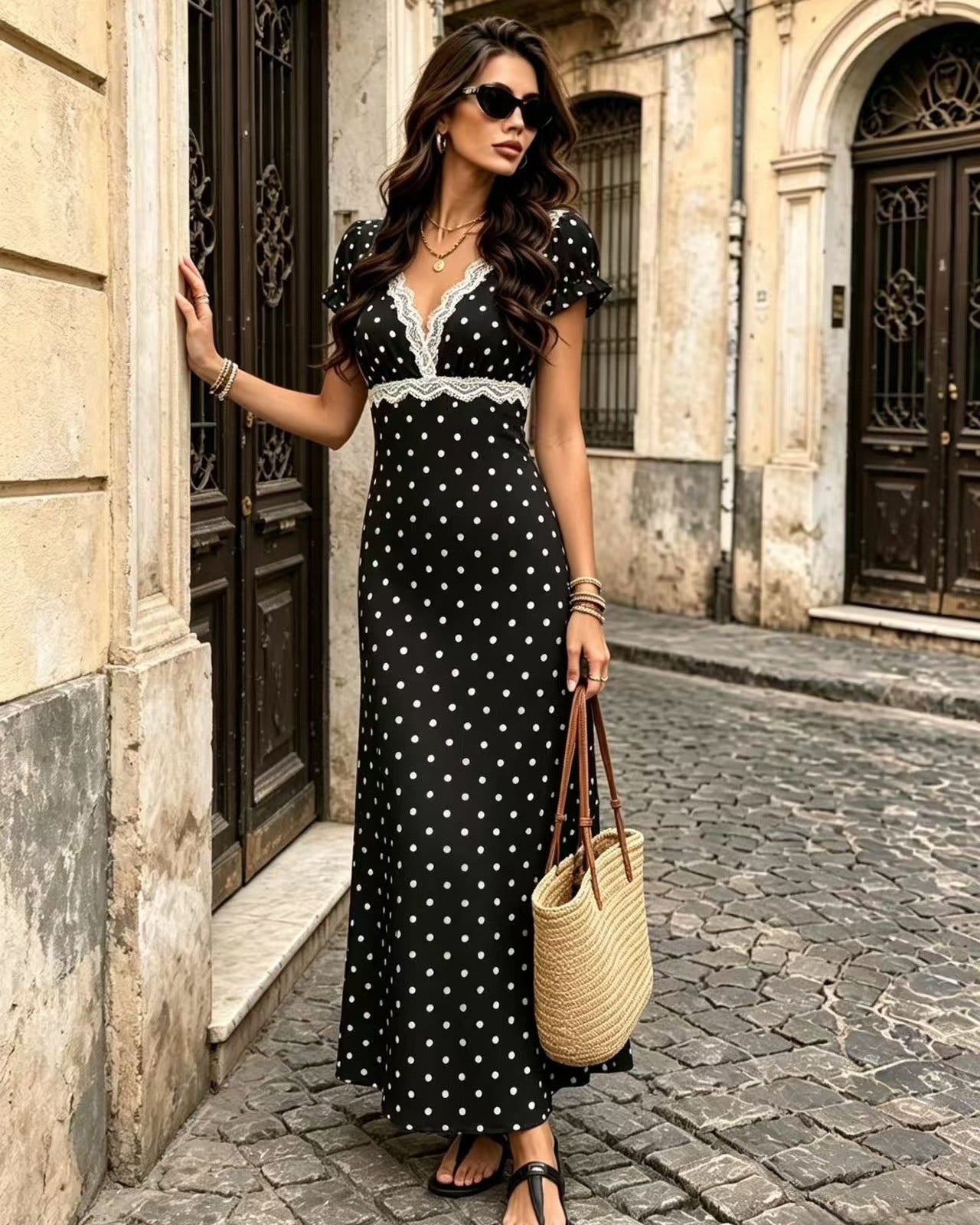 A woman stands on a cobblestone street wearing a black maxi dress with white polka dots and a white lace V-neckline. She is holding a woven straw tote bag and wearing sunglasses.