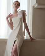 Woman in a white one-shoulder dress with a large bow standing in a room with light curtains.