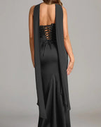A woman shown from the back wearing a sleeveless black dress with a lace-up open back and a long draped overlay, photographed against a plain light gray studio background.