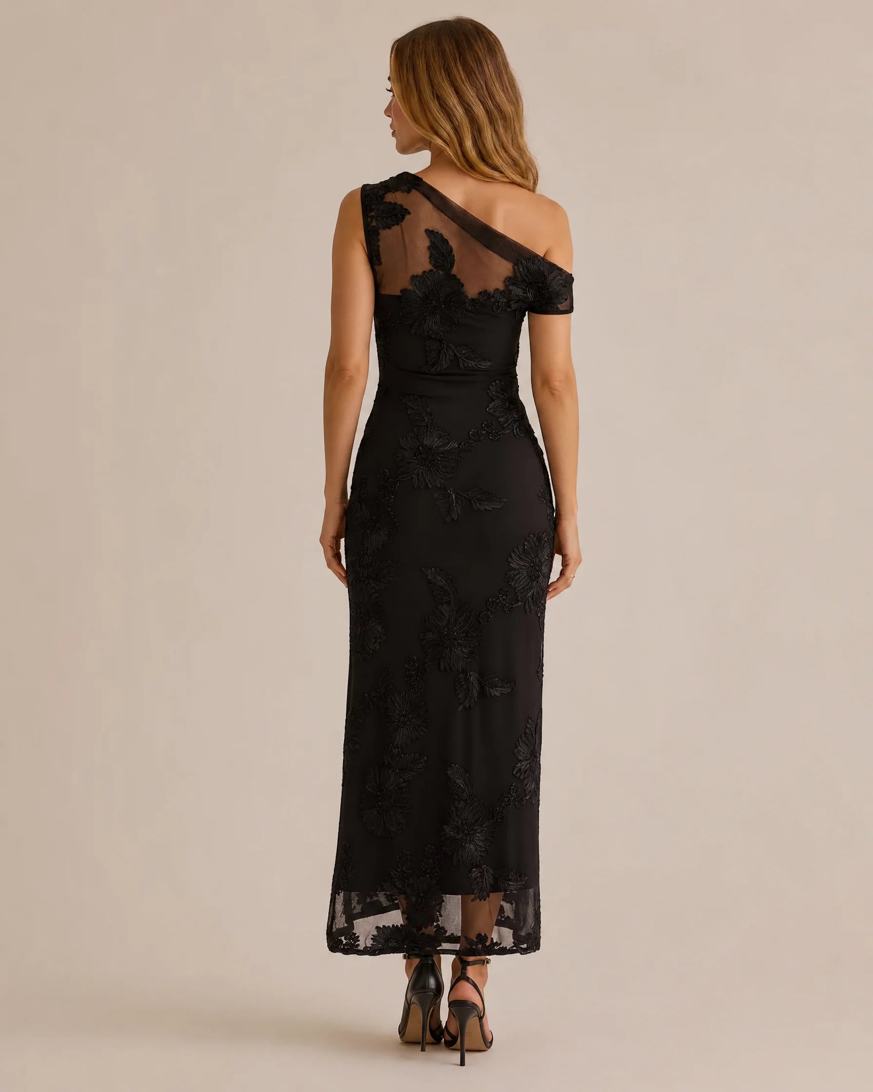 A woman modeling a black off-the-shoulder lace maxi dress from the back, shown against a plain beige studio background.