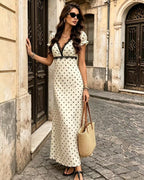 A woman stands on a cobblestone street wearing a cream maxi dress with black polka dots and a black V-neckline. She is holding a woven straw tote bag and wearing black sandals.