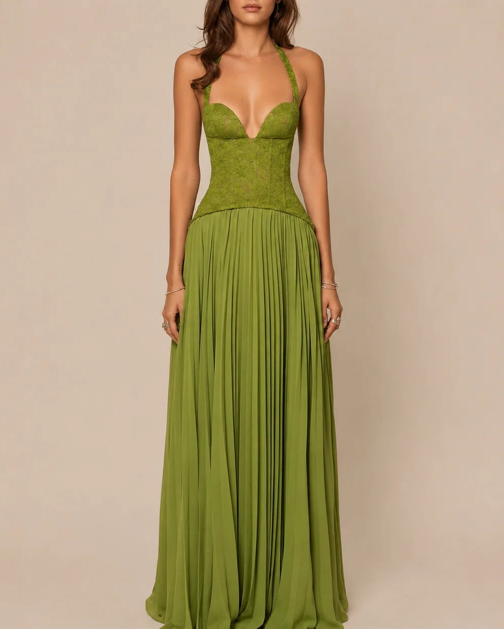 A woman modeling a floor-length olive green gown with a lace corset-style bodice, thin shoulder straps, and a flowing pleated skirt against a plain beige studio background.
