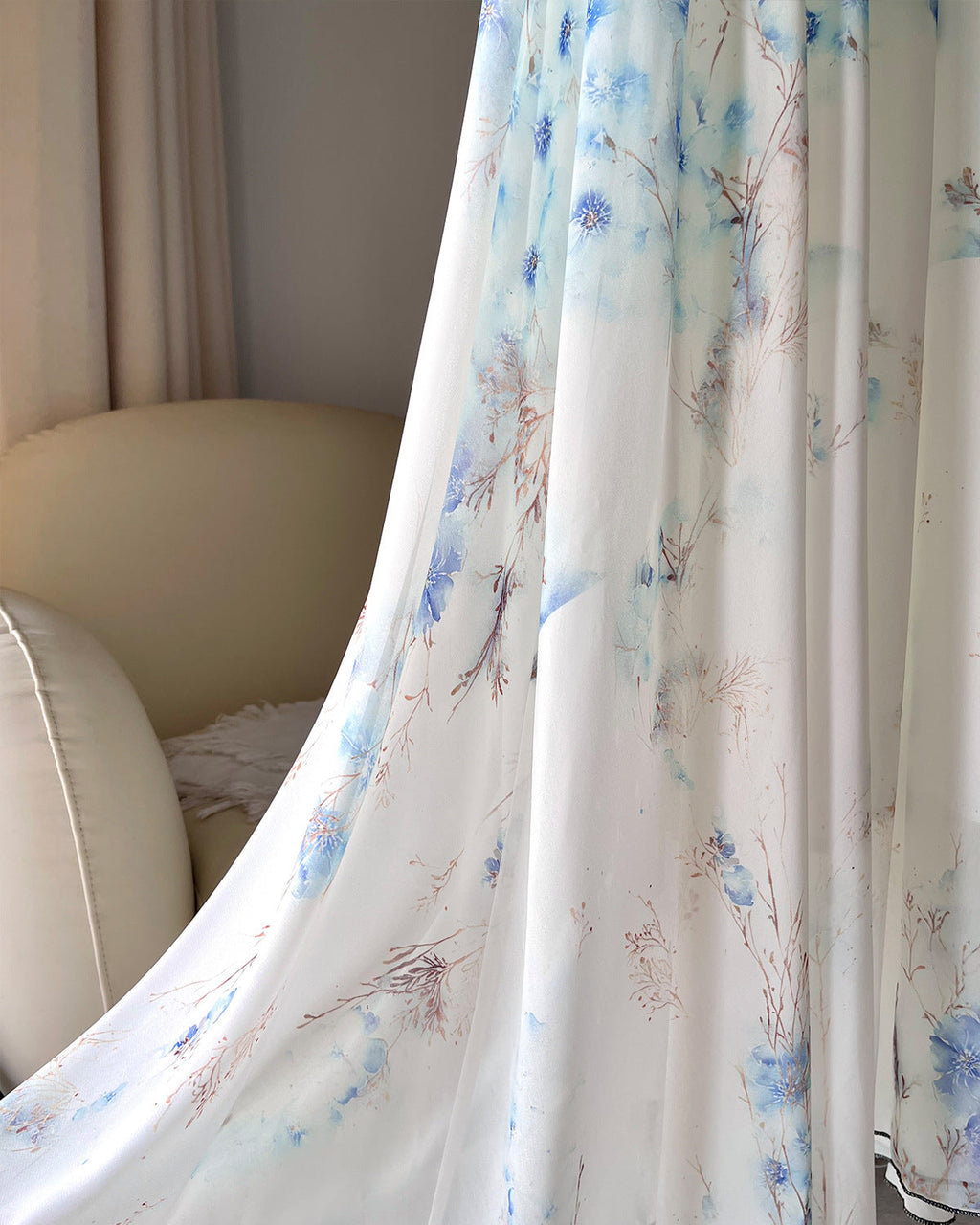 A sheer white curtain panel with a delicate blue and brown watercolor floral pattern, draped elegantly in a softly lit room next to a beige armchair.