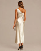 A woman shown from the back wearing a one-shoulder satin midi dress in a warm champagne or cream tone. The dress has a structured corset-style waist and a single strap across the upper back.