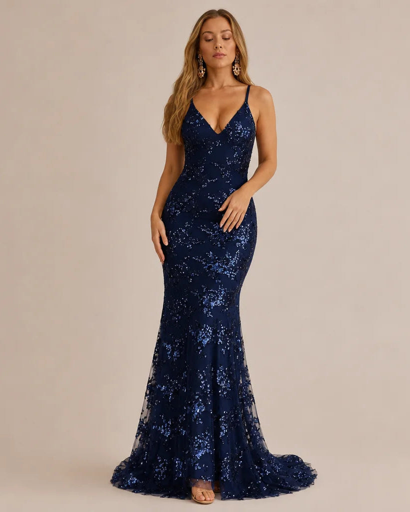 A woman modeling a navy blue sequined mermaid gown with a deep V-neckline and spaghetti straps, standing against a solid beige studio background.