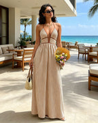 A woman stands on a resort patio wearing a beige and white vertically striped halter maxi dress with a deep V-neckline and side cutouts. She is holding a straw bag and a bouquet of flowers, with a sunny beach and ocean in the background.