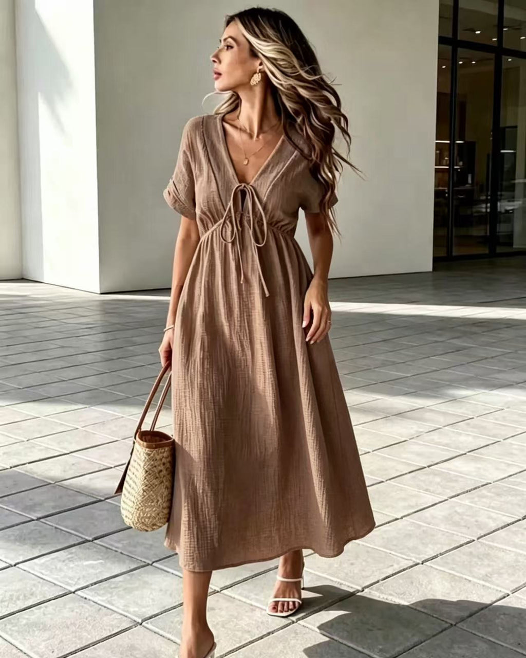A woman wearing a brown short-sleeve midi dress with a tie waist, walking outdoors on a tiled walkway while carrying a woven straw bag.