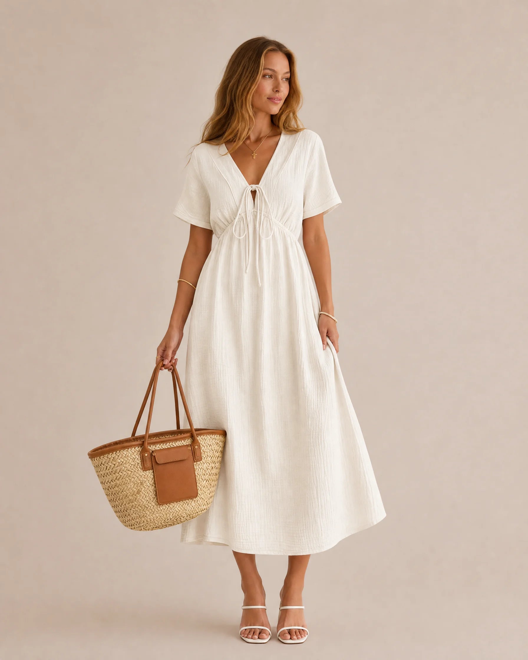 A woman modeling a short-sleeve white midi dress with a V-neckline and drawstring waist, holding a woven straw tote bag with brown handles, against a solid beige studio background.