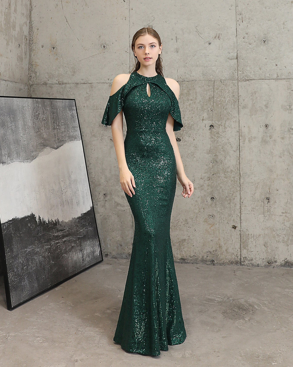 Woman in a green sequin dress standing against a concrete wall with a painting.