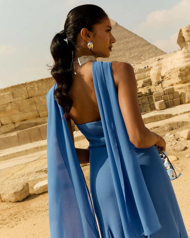 A woman photographed from behind wearing a bright blue sleeveless dress with a draped cape-style back detail, standing outdoors in front of ancient stone ruins and a pyramid under a pale sky.