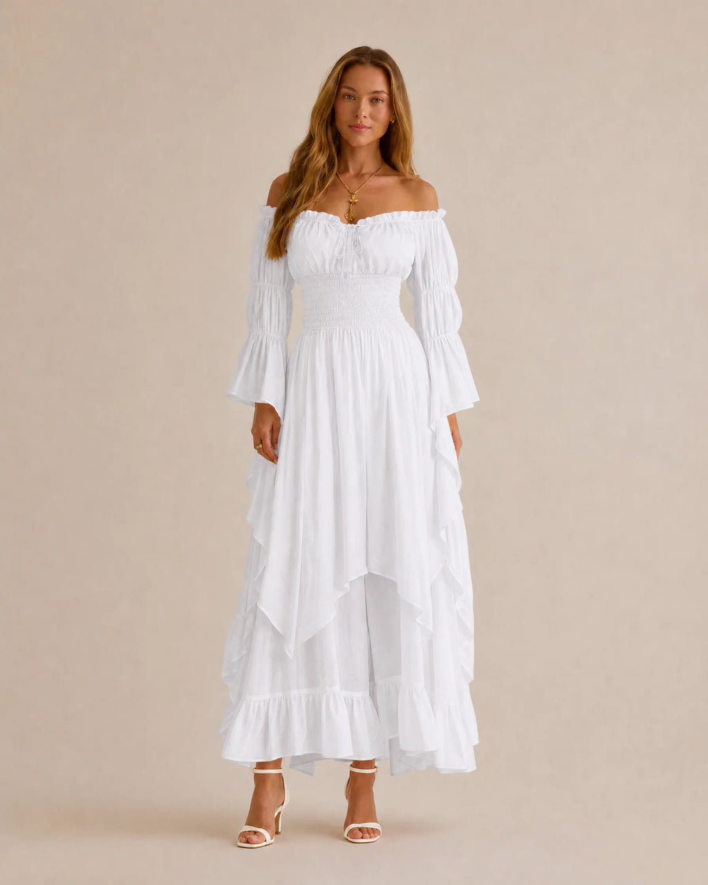 A woman modeling a white off-the-shoulder maxi dress with long flared sleeves, a smocked bodice, and a tiered ruffled skirt, photographed against a plain beige studio background.