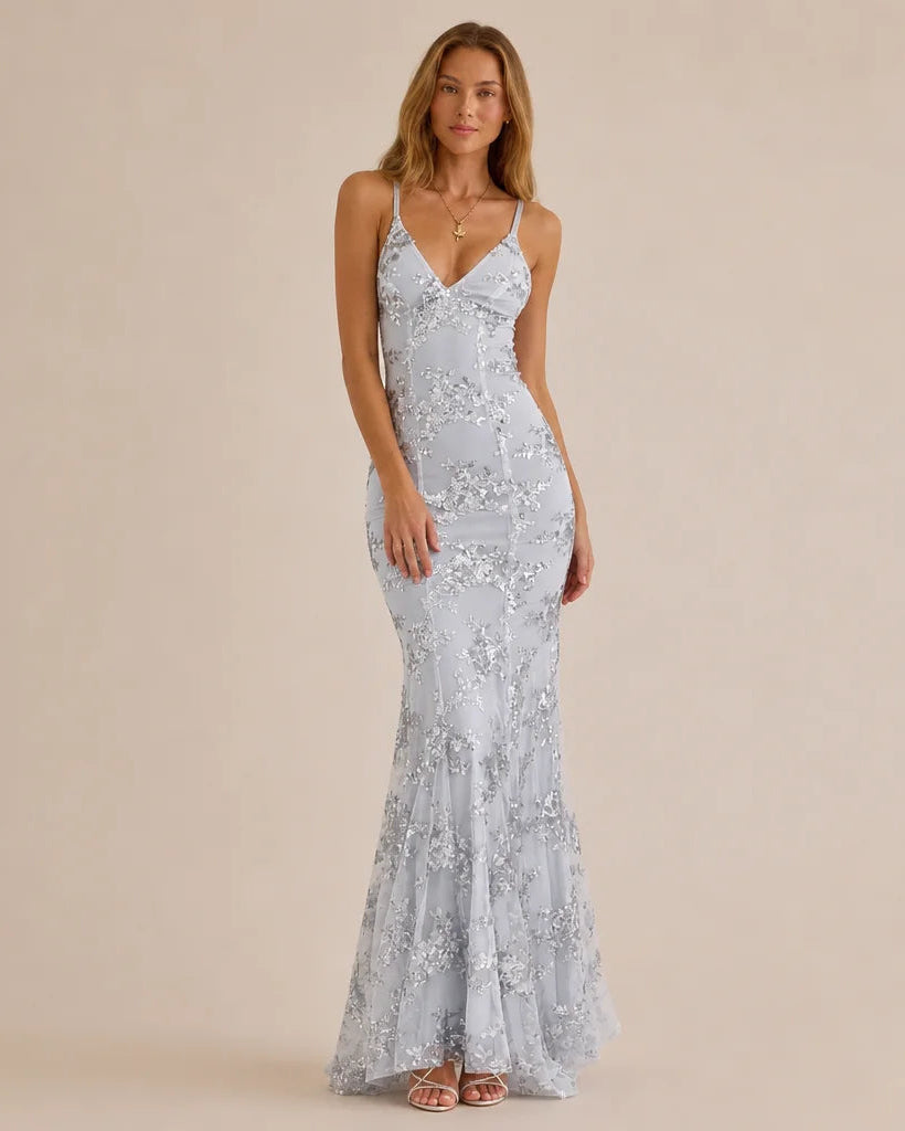 A woman modeling a floor-length light blue lace mermaid gown with thin spaghetti straps and a V-neckline, standing against a solid beige studio background.