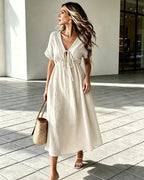A woman wearing a cream-colored short-sleeve midi dress with a tie-front detail, walking outdoors on a tiled patio while holding a woven straw bag.