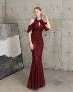 A woman models a floor-length, deep burgundy sequined gown with cold-shoulder ruffle sleeves and a keyhole cutout at the neck, standing in a concrete room next to a framed abstract print.