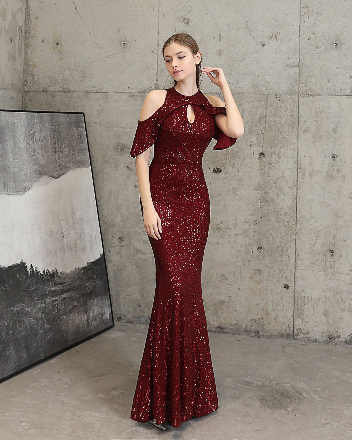 A woman models a floor-length, deep burgundy sequined gown with cold-shoulder ruffle sleeves and a keyhole cutout at the neck, standing in a concrete room next to a framed abstract print.