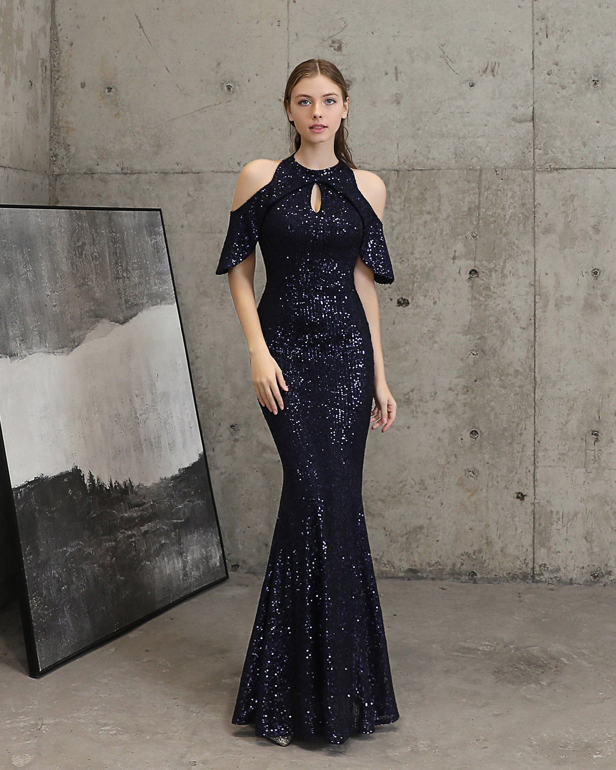 A woman stands in a studio with a concrete wall background wearing a dark navy sequined mermaid gown with cold-shoulder sleeves and a keyhole neckline. A large framed abstract print leans against the wall on the left.