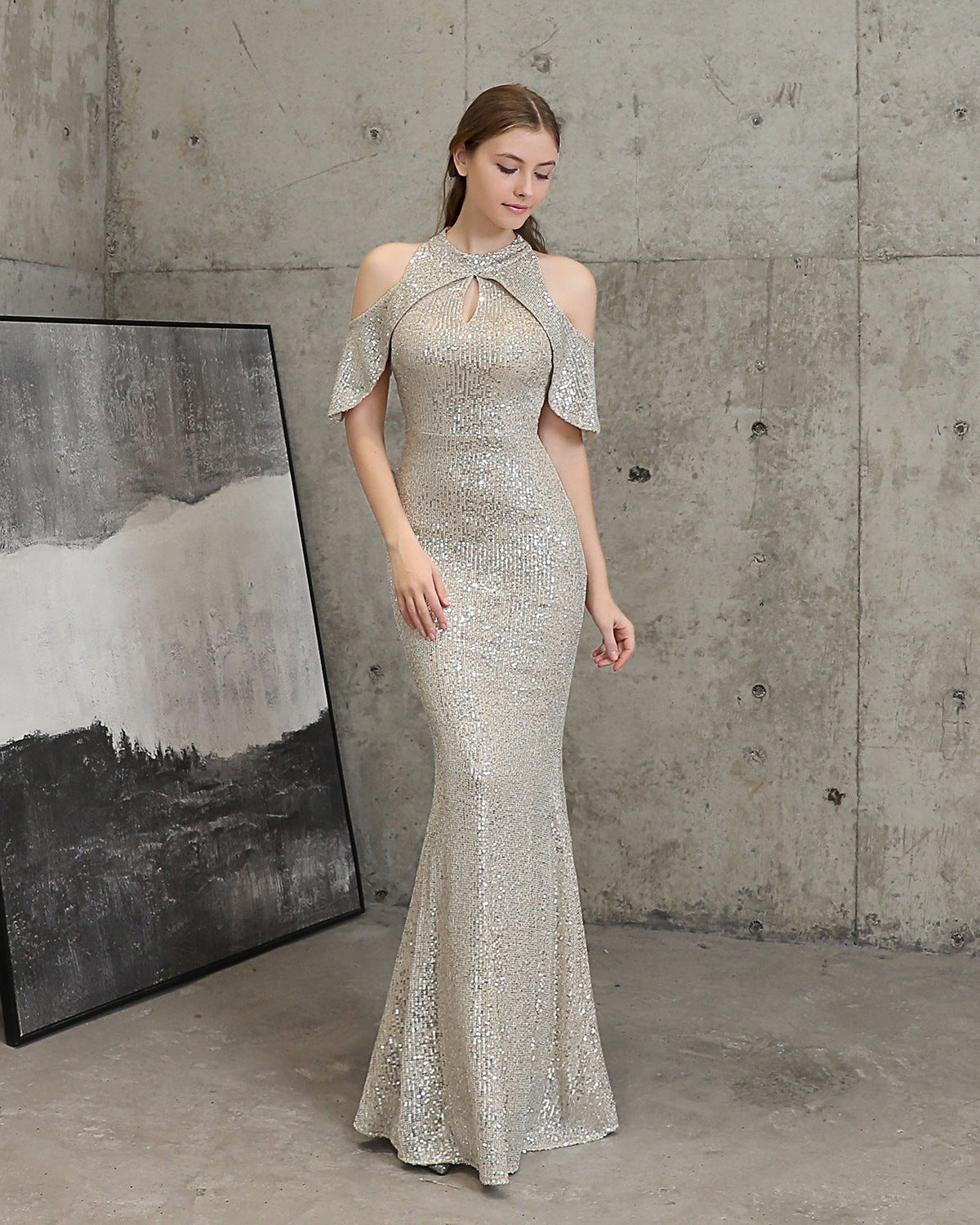 A woman models a floor-length, off-the-shoulder champagne sequin gown with a keyhole neckline and flared cuffs, standing in a concrete room next to a framed abstract print.