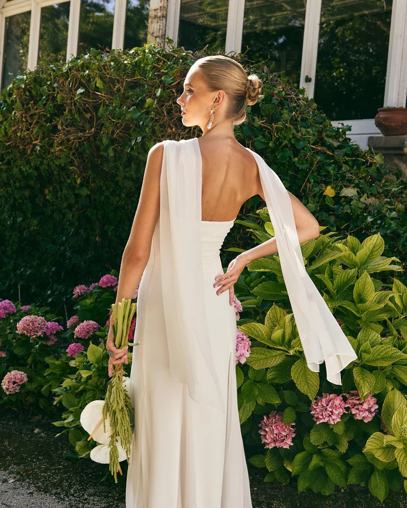 Woman in a white dress with a cape standing in a garden with pink flowers and greenery.