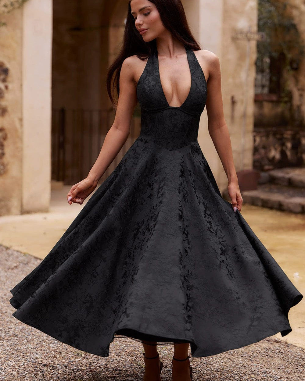 A woman wearing a black lace halter midi dress with a deep V-neckline and flared skirt, standing outdoors in front of a stone archway.