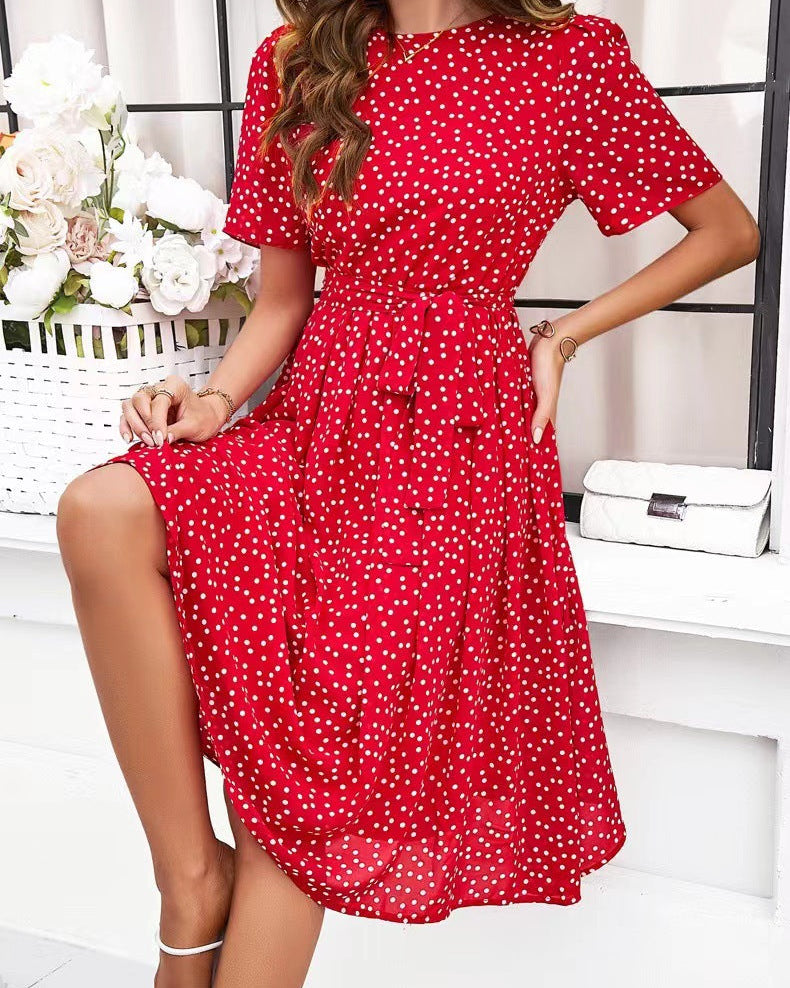 Red polka dot dress worn by a model in a minimalistic setting with flowers and a handbag.