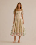 A woman modeling a cream-colored midi dress with a delicate floral and vine pattern, featuring short flutter sleeves and a square neckline, set against a solid beige studio background.