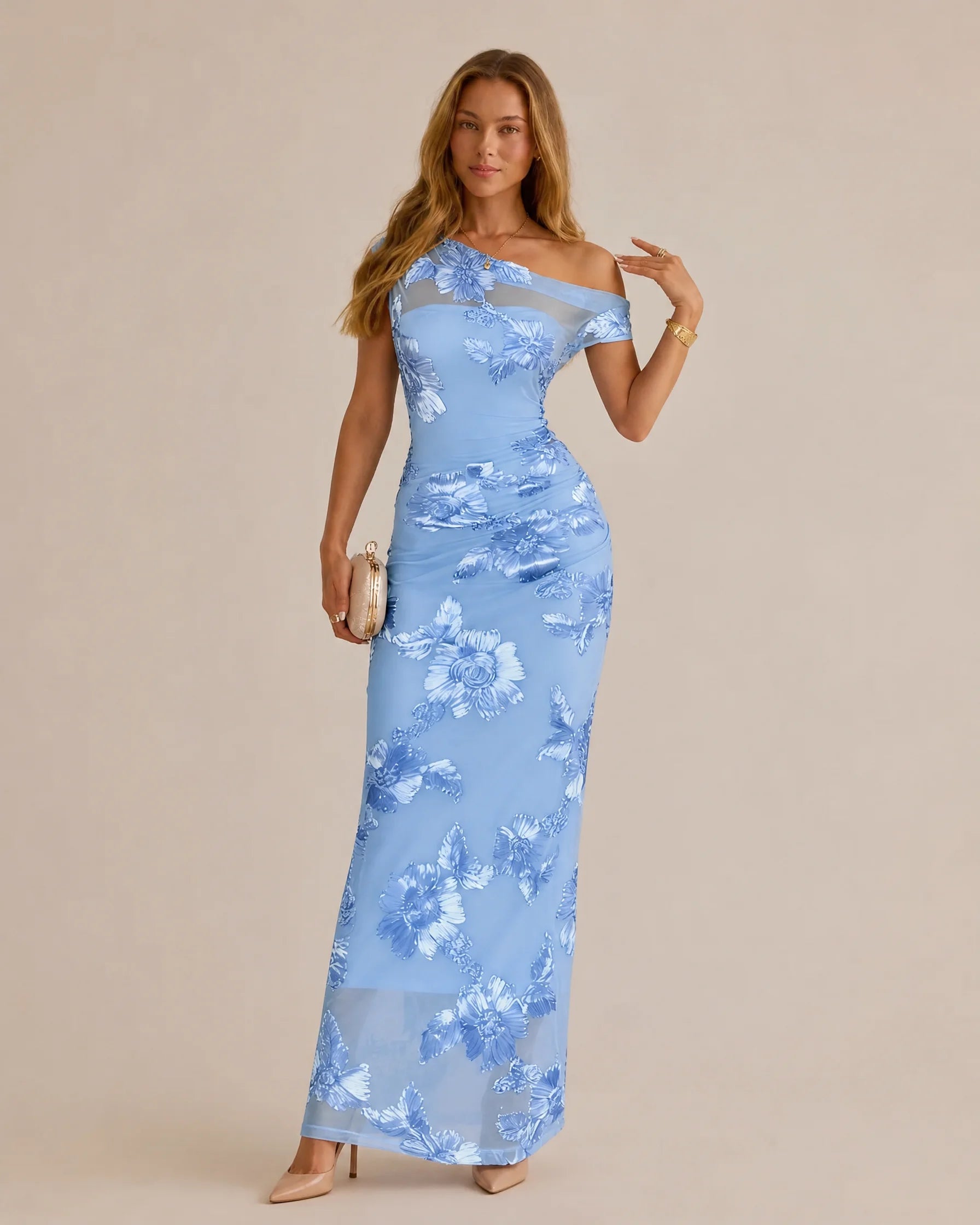 A woman models a light blue off-the-shoulder maxi dress with a white floral pattern, standing against a solid beige studio background.