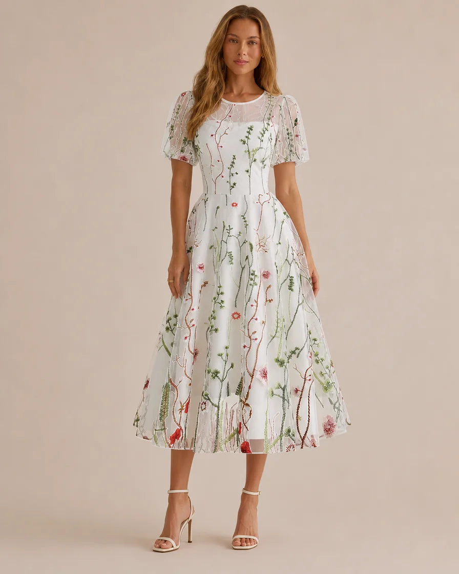 A woman modeling a white midi dress with a delicate green and red botanical print, sheer puff sleeves, and a round neckline, set against a solid beige studio background.