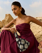 A woman outdoors in a desert-like rocky setting wearing a strapless deep burgundy dress and holding a black handbag with large white floral embroidery.