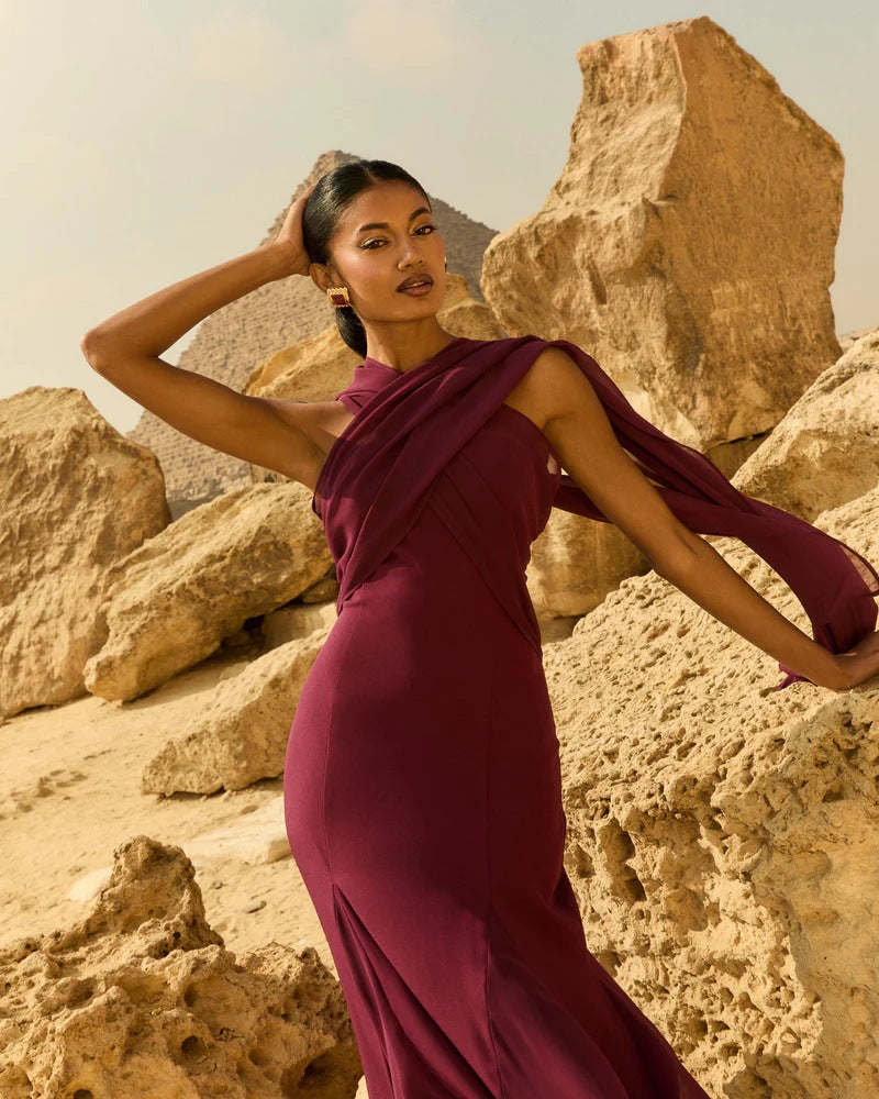 A woman modeling a sleeveless, draped magenta-purple maxi dress in a sandy desert landscape with large rock formations.