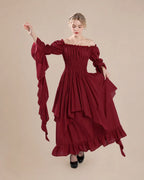 A woman modeling a long off-the-shoulder dress in deep burgundy. The dress features a smocked bodice, voluminous long sleeves with dramatic cascading ruffles, and a layered, tiered skirt with a ruffled hem.