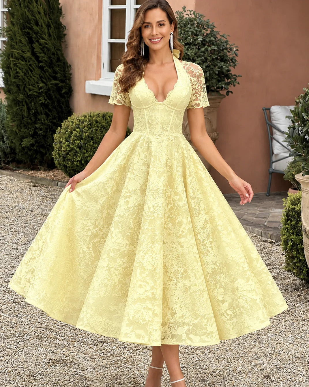 A woman wearing a pale yellow lace A-line dress with short lace sleeves, a deep V-neckline, and a full skirt, standing outdoors on a gravel path in front of a stucco building.