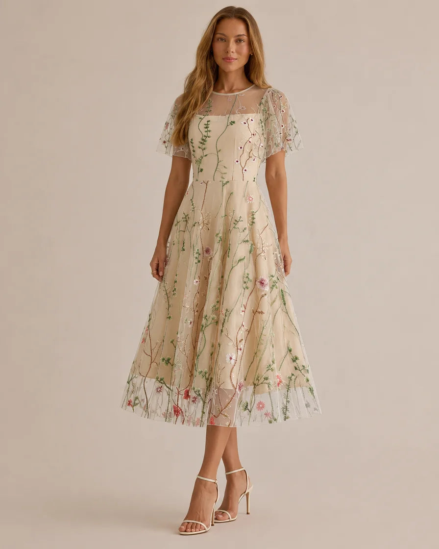 A woman models a beige midi dress with delicate floral embroidery and sheer flutter sleeves against a solid beige studio background.