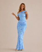 A woman modeling a light blue off-the-shoulder maxi dress with a fitted silhouette and a floral lace overlay, posed against a solid beige studio background.