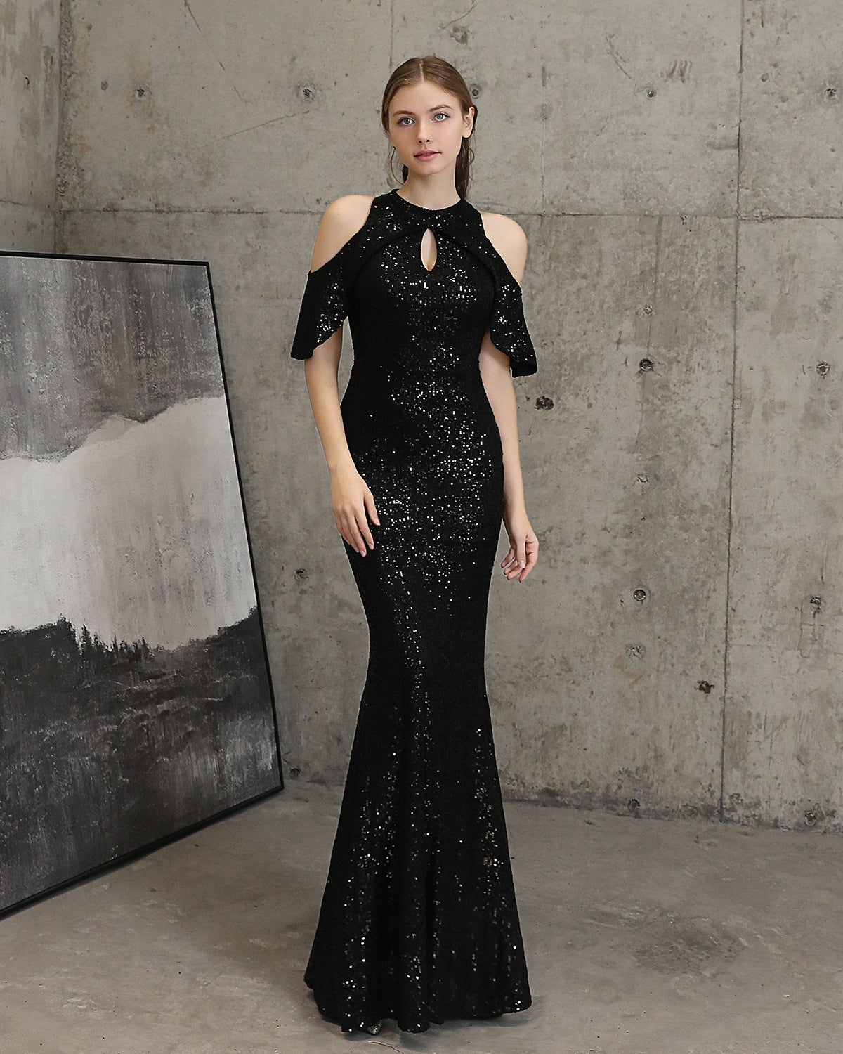 A woman stands in a studio with a concrete wall and a large framed black-and-white abstract painting, wearing a fitted black evening gown covered in sequins. The dress has cold-shoulder short sleeves, a keyhole neckline, and a mermaid-style flared hem.