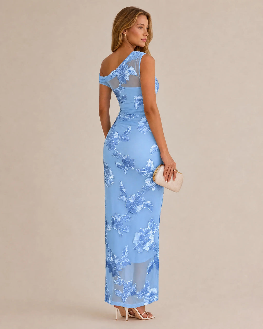 A woman modeling a light blue one-shoulder maxi dress with a floral lace pattern, shown from a three-quarters back view against a solid beige background.