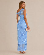 A woman modeling a light blue one-shoulder maxi dress with a floral lace pattern, shown from a three-quarters back view against a solid beige background.