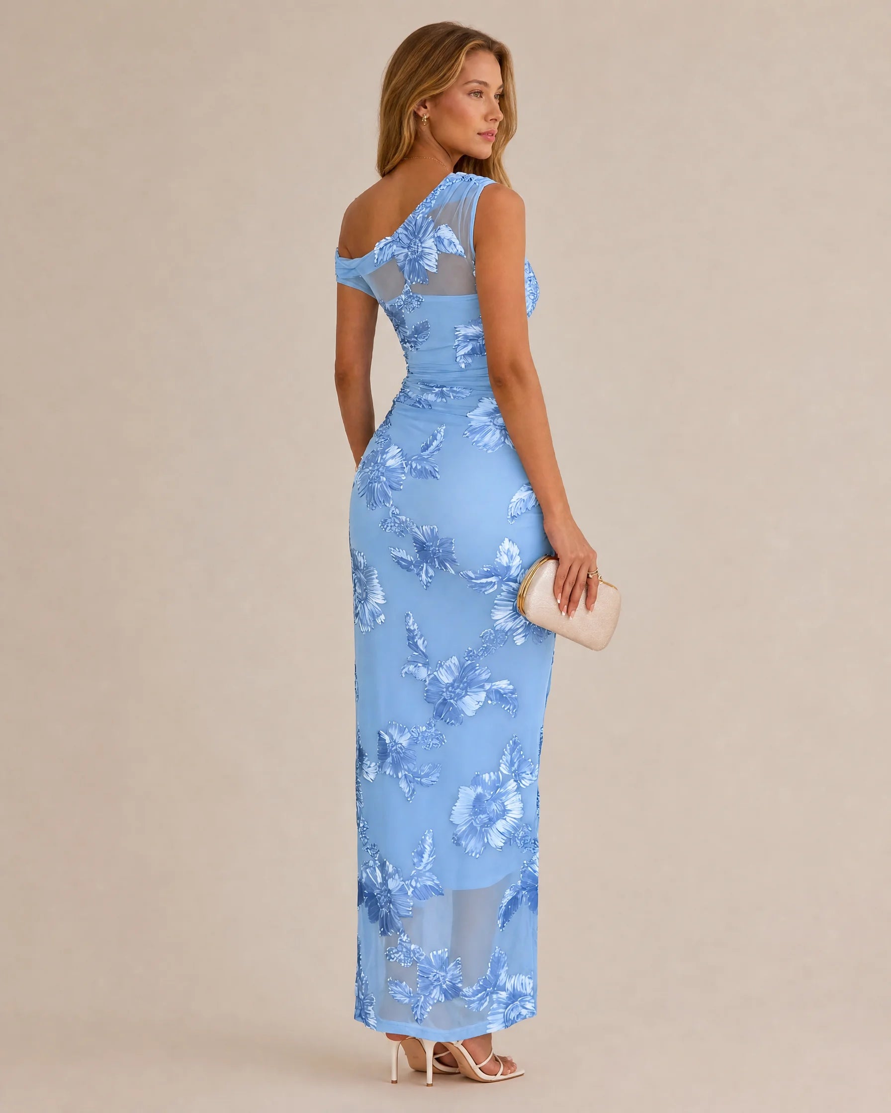 A woman modeling a light blue one-shoulder maxi dress with a floral lace pattern, shown from a three-quarters back view against a solid beige background.